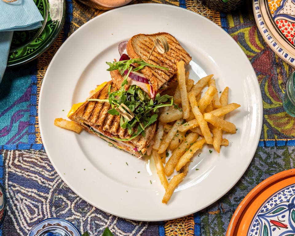 Grilled sandwich with arugula, red onion, and fries on a white plate with a colorful tablecloth.