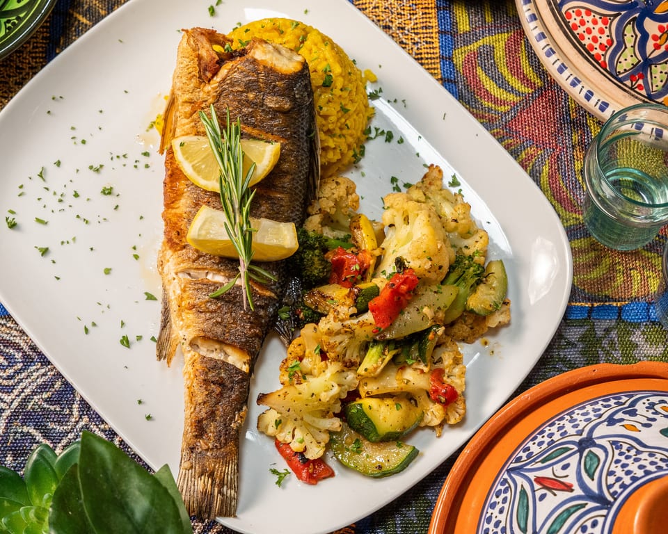 Whole roasted fish with lemon, rosemary, yellow rice, and roasted vegetables on a white plate.