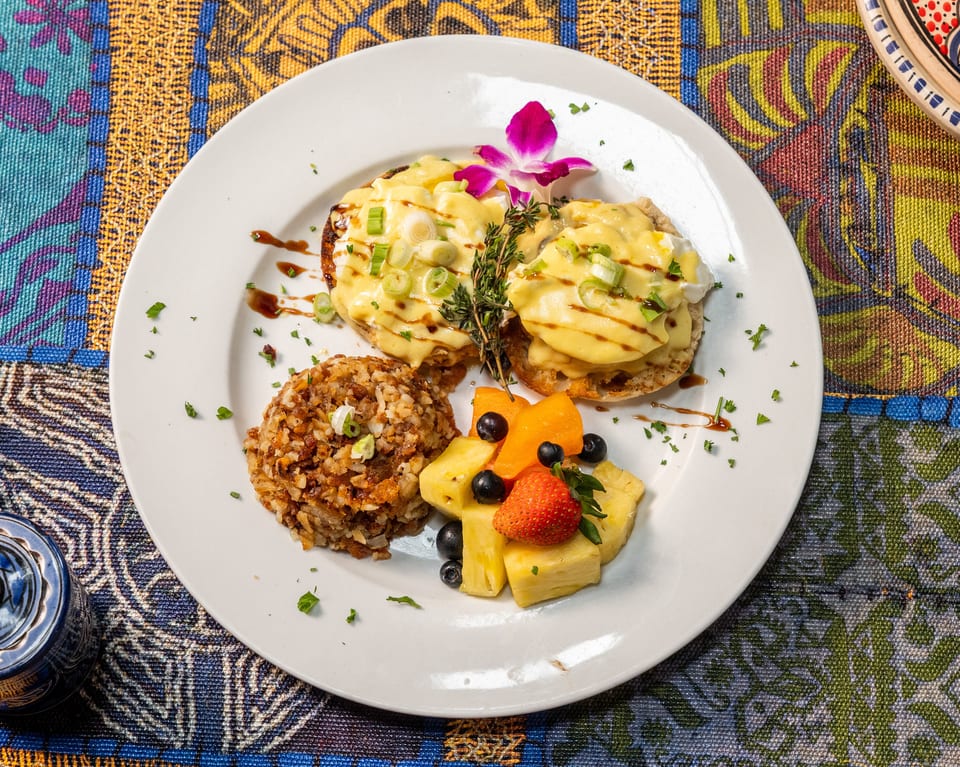 Eggs Benedict with rice and fruit salad, garnished with an orchid, on a colorful patterned tablecloth.