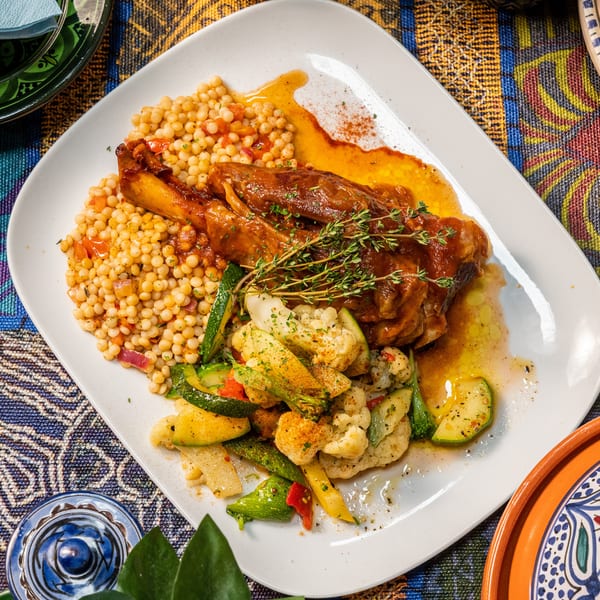 Braised lamb shank with couscous, roasted vegetables, and fresh thyme on a white plate.