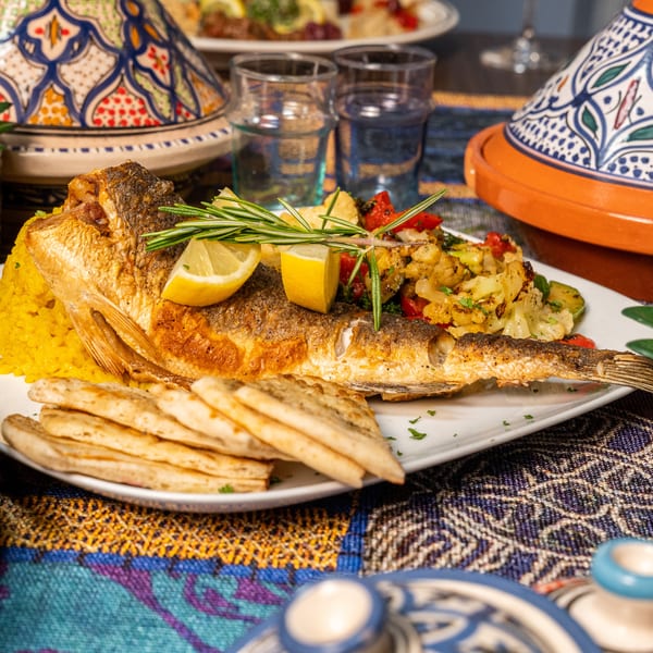 Whole fried fish with yellow rice, roasted vegetables, lemon, rosemary, and flatbread on a patterned tablecloth.