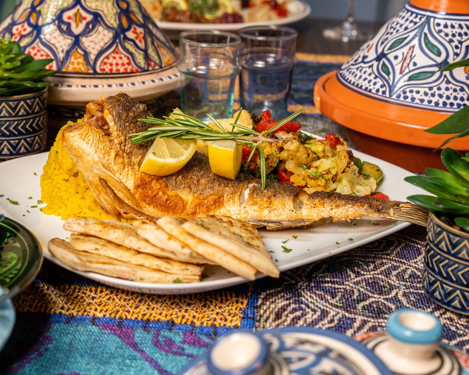 Whole fried fish with yellow rice, roasted vegetables, lemon, rosemary, and flatbread on a patterned tablecloth.