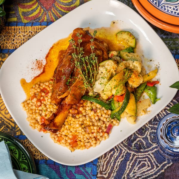 Braised lamb shank with couscous, roasted vegetables, and fresh thyme on a white plate.