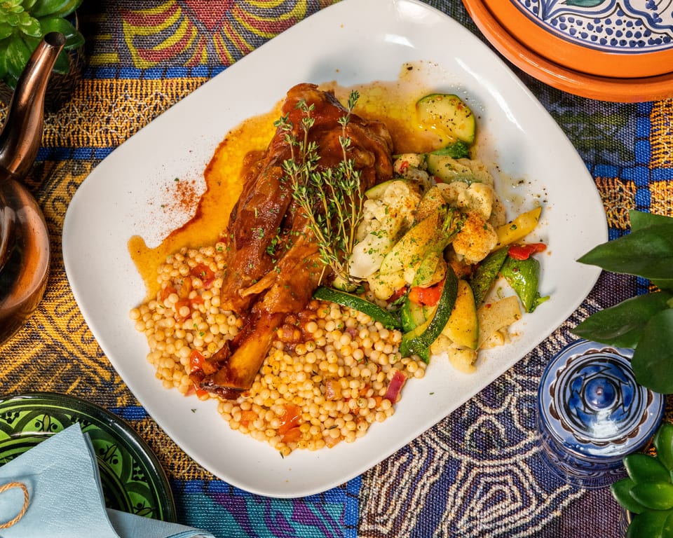 Braised lamb shank with couscous, roasted vegetables, and fresh thyme on a white plate.