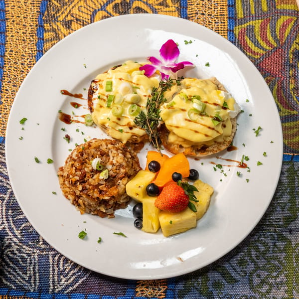 Eggs Benedict with rice and fruit salad, garnished with an orchid, on a colorful patterned tablecloth.