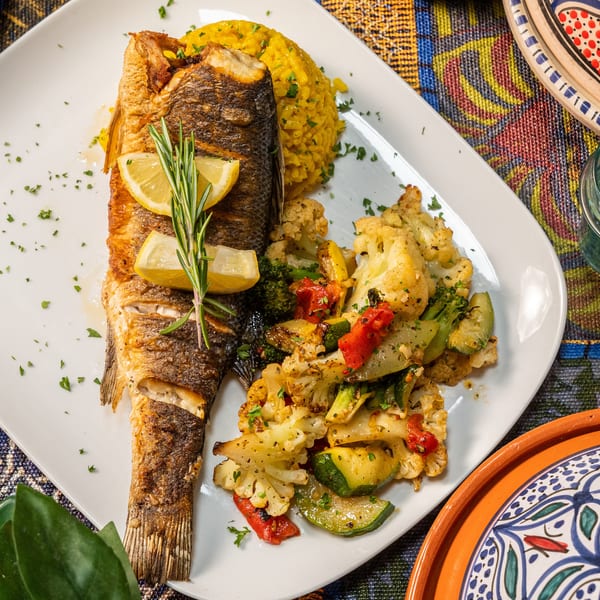 Whole roasted fish with lemon, rosemary, yellow rice, and roasted vegetables on a white plate.