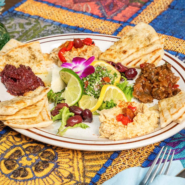 A colorful appetizer platter with various dips, pita bread, olives, and lemon slices on a patterned tablecloth.