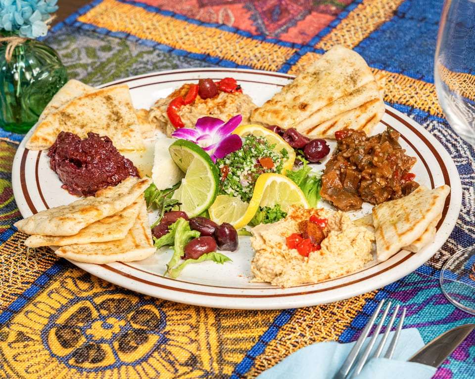 A colorful appetizer platter with various dips, pita bread, olives, and lemon slices on a patterned tablecloth.